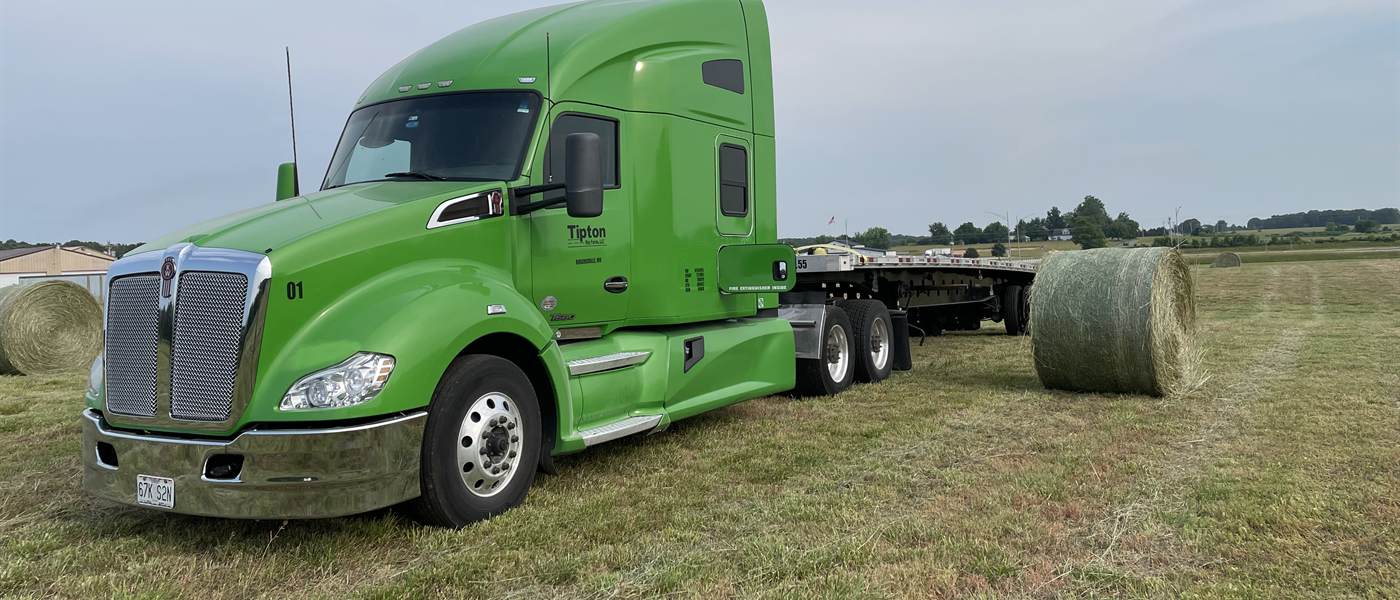 A green Tipton Hay Farms Kenworth on a flatbed, loading round bales off a hay field